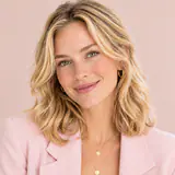 Selena, blonde aux cheveux ondulés, sourit face à l’objectif, en blazer rose poudré sur fond rose clair.